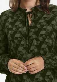 Blouse verte à motifs avec un col à volants et une fermeture à nouer. Comprend des formes géométriques et des manches texturées pour plus de détails.