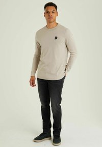 Beige Langarm-Shirt mit Rundhalsausschnitt und kleinem schwarzen Logo-Patch, kombiniert mit dunklen Jeans und grünen Schuhen, stehend vor einem hellen Hintergrund.