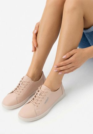 Beine an den Knöcheln gekreuzt, tragen hellrosa Schnürschuhe mit "VENEZA"-Logo, Hände ruhen sanft auf den unteren Beinen, sitzend auf weißem Boden.