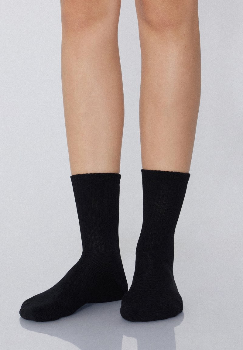 Czarne bawełniane skarpetki za kostkę z żeberkowym mankietem, dopasowanym krojem i gładką teksturą, spoczywające na nagich nogach na neutralnym tle.