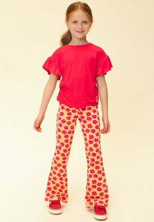 Jong meisje dat glimlacht, draagt een felrood shirt met ruches aan de mouwen, gele broek met rood bloemenpatroon en rode sneakers, staand tegen een lichte achtergrond.