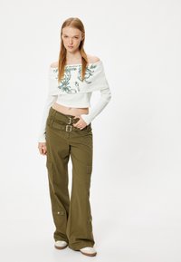 Weißes, schulterfreies, geripptes Cropped-Top mit grünem Grafikdruck, kombiniert mit lockeren, olivgrünen Cargo-Hosen mit mehreren Taschen und einem Schnallengürtel.
