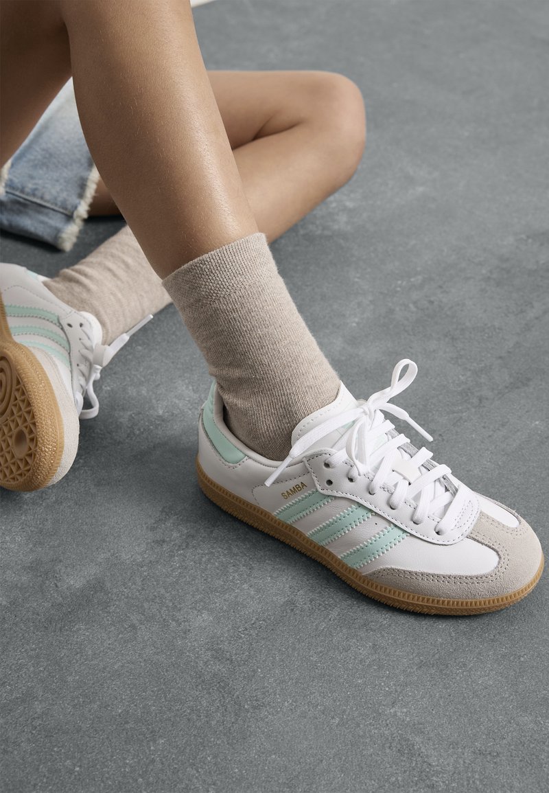 Zapatillas deportivas blancas con rayas verde menta y acentos beige, que cuentan con una suela texturizada y un upper suave, combinadas con calcetines beige.