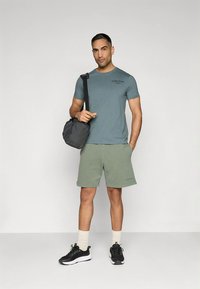 Lys blå t-skjorte med rund hals for menn med svart trykk, grønne shorts, svarte joggesko og en grå veske, stående mot en ensfarget bakgrunn.