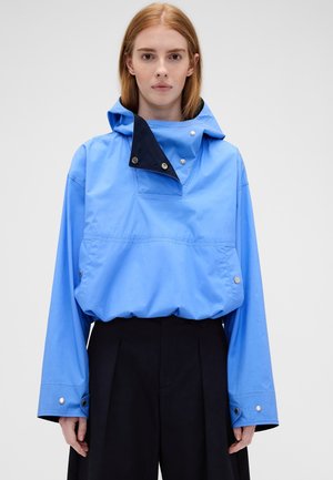 Femme portant une veste à capuche bleue avec doublure intérieure noire et pantalon noir à jambes larges sur un fond clair uni.