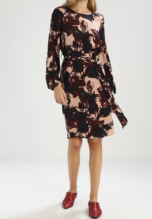 Femme portant une robe beige mi-longue aux genoux avec des motifs floraux noirs et rouge foncé, manches longues, ceinture à la taille et chaussures rouges pointues.