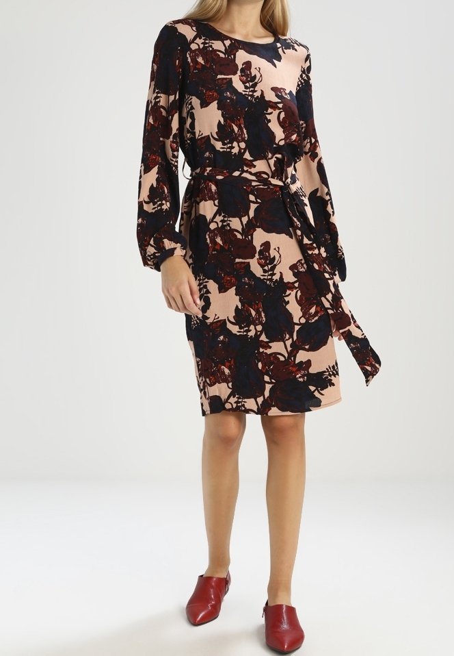 Femme portant une robe beige mi-longue aux genoux avec des motifs floraux noirs et rouge foncé, manches longues, ceinture à la taille et chaussures rouges pointues.