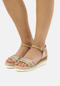 Las sandalias de cuña presentan tiras trenzadas en beige, morado y verde, con una puntera redondeada y una correa en el tobillo. La suela de corcho y goma proporciona estabilidad.