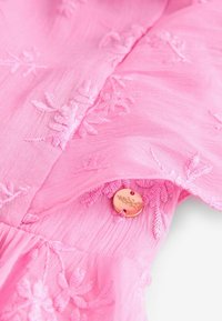 Roze geborduurde stof met bloemmotieven en een ronde metalen knop met bloemengraving, die textuur en detail toevoegt.