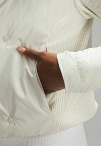 Lekka, kremowa kurtka o gładkiej powierzchni, z żeberkowanymi akcentami i detalami guzików; zawiera boczną kieszeń oraz wytłoczone logo na mankiecie.