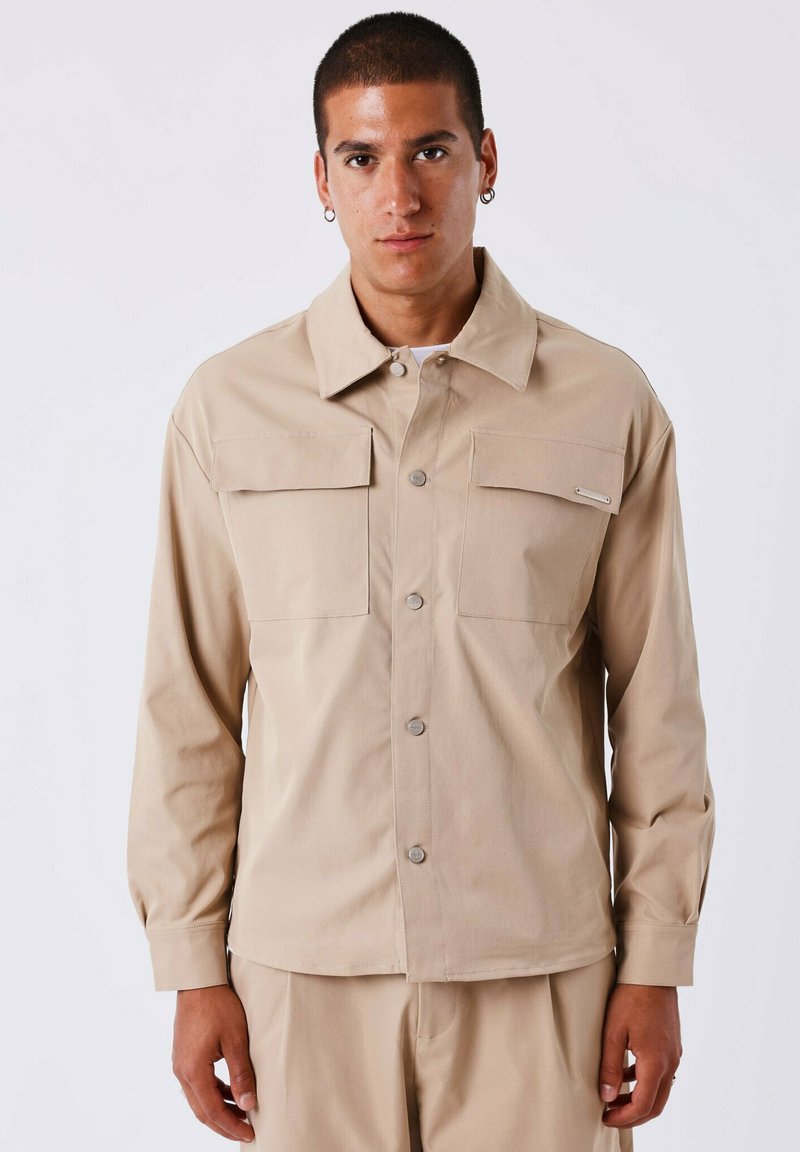 Camicia beige a maniche lunghe con due tasche sul petto e chiusura a bottoni. Realizzata in tessuto morbido con colletto e polsini, presenta dettagli in metallo argentato.