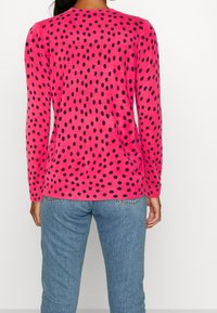 Maglione fucsia con pois blu navy, caratterizzato da un colletto rotondo e polsini a coste. Indossato con jeans azzurri, che mostrano una vestibilità comoda.