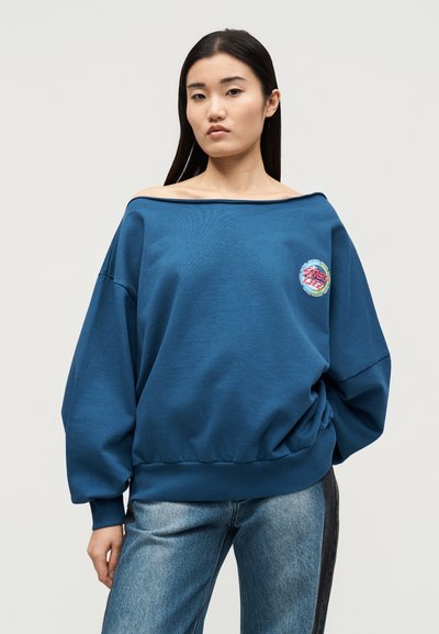 Νεαρή γυναίκα που φοράει χαλαρό μπλε φούτερ τύπου off-shoulder με πολύχρωμο σχέδιο και δίχρωμο μπλε τζιν, στέκεται μπροστά σε απλό φόντο.