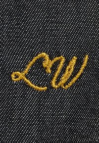 Kullast niidist tikand südame ja stiliseeritud "W" mustal teksariimise kangal.