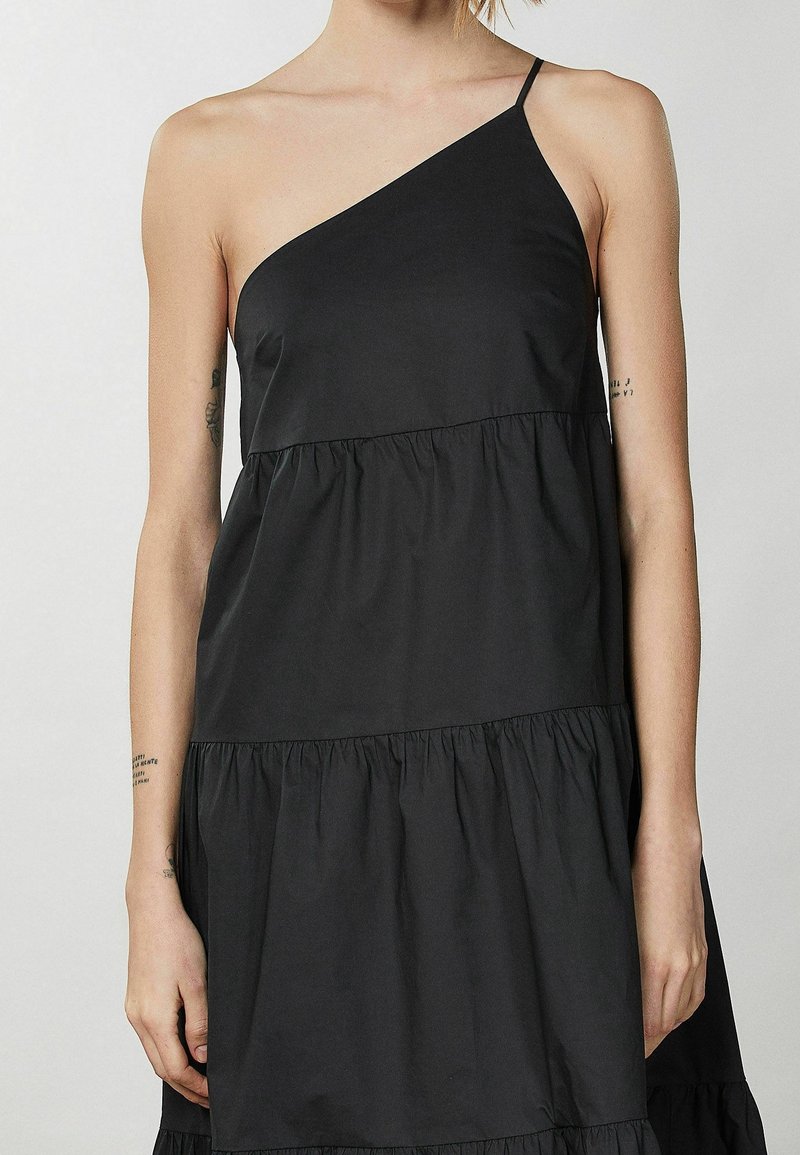 Vestido negro con diseño de un solo hombro, tela en capas y textura ligera. Presenta un corte holgado y una tira delgada en el hombro.