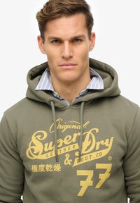 Oliven grøn hættetrøje med gult trykt logo og tekst. Materiale i bomuldsblanding med snore og et lagdelt look.