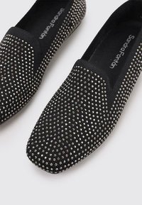 Zapatos negros tipo slip-on con una superficie de tela suave, adornados con rhinestones plateados. Presentan una suela plana y un diseño de punta redonda.