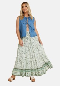 Denim-vesti, jossa on solmittu etuosa, yhdistettynä valkoiseen virtaavaan, kerrokselliseen hameeseen, jossa on vihreitä kukkakuva-aiheita ja kuvioitu helma. Ruskeat korokepohjaiset sandaalit.