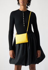 Robe noire à rayures côtelées à manches longues avec un col rond et des détails de boutons, associée à un sac bandoulière en cuir jaune.
