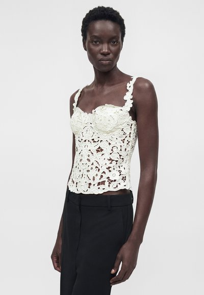 Γυναίκα με λευκό αμάνικο τοπ από δαντέλα με λουλούδια και μαύρο παντελόνι, στέκεται μπροστά σε ένα απλό ανοιχτό γκρι φόντο.