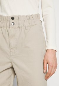 Pantalon beige taille haute avec ceinture élastique et deux boutons-pression noirs, porté avec un haut blanc à manches longues côtelé, main reposant le long du corps.