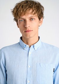 Camisa de botones azul claro hecha de tejido texturizado; cuenta con un cuello, un bolsillo frontal y botones blancos. Diseño simple con detalles sutiles.