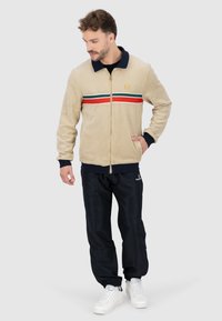 Beige Zip-Jacke mit marineblauem Kragen, die rote, grüne und weiße horizontale Streifen aufweist. Kombiniert mit schwarzen Hosen und weißen Turnschuhen.