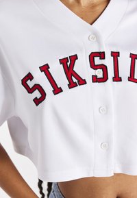 Nahaufnahme einer Person, die ein weißes, kurz geschnittenes, geknöpftes Hemd trägt, mit "SIKSILK" in großen roten Buchstaben über der Brust.