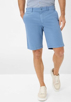 Shorts en coton bleu avec une ceinture plate, des passants de ceinture, des poches latérales et une coupe décontractée, associés à des baskets beiges et une chemise à motifs.