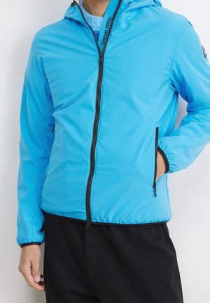 Person trägt eine leuchtend blaue Kapuzenjacke mit schwarzem Reißverschluss und schwarze Hose, Hand in der Jackentasche, vor einem schlichten hellen Hintergrund.