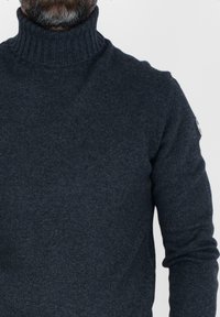 Maglione a collo alto di un profondo colore blu navy, realizzato in tessuto a costine, con una vestibilità aderente e colletto arrotolato, completo di una piccola toppa con logo sulla manica.