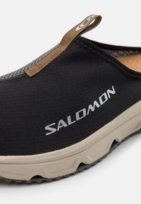 Chaussure noire à enfiler avec un dessus texturé, tirette d'accent marron, et une semelle en caoutchouc beige dotée d'un motif pour une meilleure traction. Marque bordée de sel.