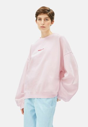 Roze oversized sweatshirt met afgezakte schouders en geribbelde manchetten, voorzien van kleine rode geborduurde tekst vlakbij de halslijn. Gecombineerd met lichtblauwe spijkerbroek.