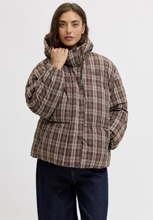 Femme portant une veste matelassée à carreaux marron et beige avec un col montant, posant avec une main près du menton et l'autre dans une poche.