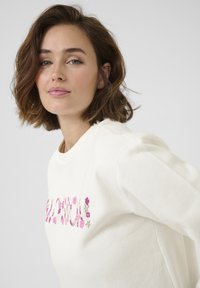 Cream sweatshirt featuring floral embroidery in pink and beige on the front. The fabric has a soft texture and relaxed fit.
