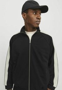 Sort zip-jakke med lyse sidelister, høj krave og elastiske ærmer. Stoffet ser glat ud med en mat finish.