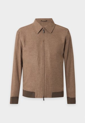Homme portant une veste marron, une chemise rayée, un pantalon crème et des chaussures marron, tenant un sac beige avec des garnitures en cuir marron, debout devant un fond uni.