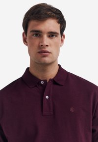 Polo shirt bordeaux con colletto, pattina a due bottoni e logo ad albero ricamato sul lato sinistro del petto; materiale in cotone con finitura testurizzata.