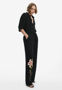 Combinaison noire à manches courtes, avec un motif floral sur la jambe gauche. Tissu doux, coupe ample, comprend une ceinture et des poches.
