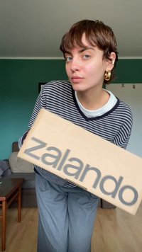 Brown cardboard box with "zalando" printed in large gray letters. Striped sweater and gray pants worn by person holding the box.