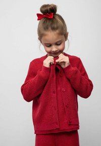 Czerwony sweter na guziki z teksturowanym wzorem, z przodu z guzikami i ściągaczami przy mankietach. Pod spodem noszona jest dopasowana czerwona bluzka.