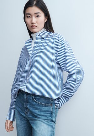 Žena s dlhými tmavými vlasmi, ktorá má na sebe modro-bielu pruhovanú košeľu na gombíky prekrytú bielym rolákom a modré džínsy, stojaca pred svetlým pozadím.