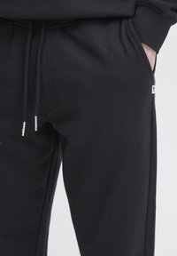 Schwarze Sweatpants aus weichem Stoff mit einem Kordelzugbund, ausgestattet mit Seitentaschen und einem kleinen Logoschild auf der linken Seite.
