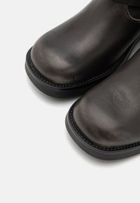 Saboti din piele maro cu vârf rotunjit, textură netedă și o strălucire subtilă. Talpa din cauciuc negru cu detalii cusute de-a lungul marginii.