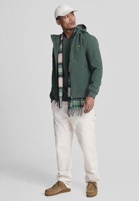 Groene ritsjas, zwarte trui, crèmekleurige broek en bruine schoenen. Geaccentueerd met een gestreepte sjaal en een witte pet, met een logo op de jas.