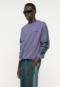 Purple sweatshirt with a round neck, ribbed cuffs, and blue striped accents along the sleeves, paired with green sweatpants featuring side stripes.