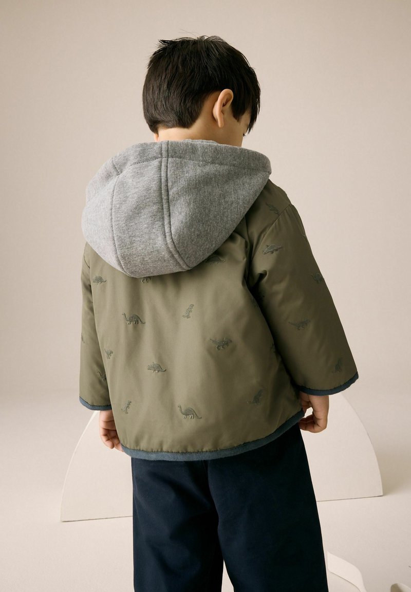 Enfant de dos portant une veste verte avec des motifs de dinosaures et une capuche grise, debout devant un fond beige uni.