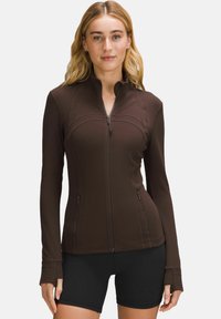 Brown zip-up athletic jacket with long sleeves and thumbholes, featuring seam accents and two zippered pockets on the front.