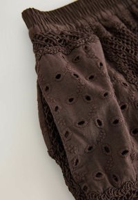 Brown fabric with embroidered eyelet patterns and crocheted lace detailing on a garment sleeve or pant leg.