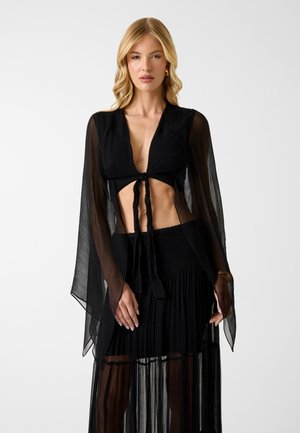 Blonde woman wearing a sheer black tie-front top with long bell sleeves and a matching sheer pleated skirt, standing against a white background.